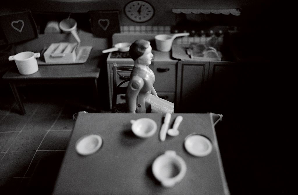 A female doll figure sits in the miniature kitchen of a dollhouse. She is positioned between a set table in the foreground and a cluttered kitchen unit in the background. She sits turned to the side and appears to be looking out of a window through which light falls. The photograph is rendered in black and white.