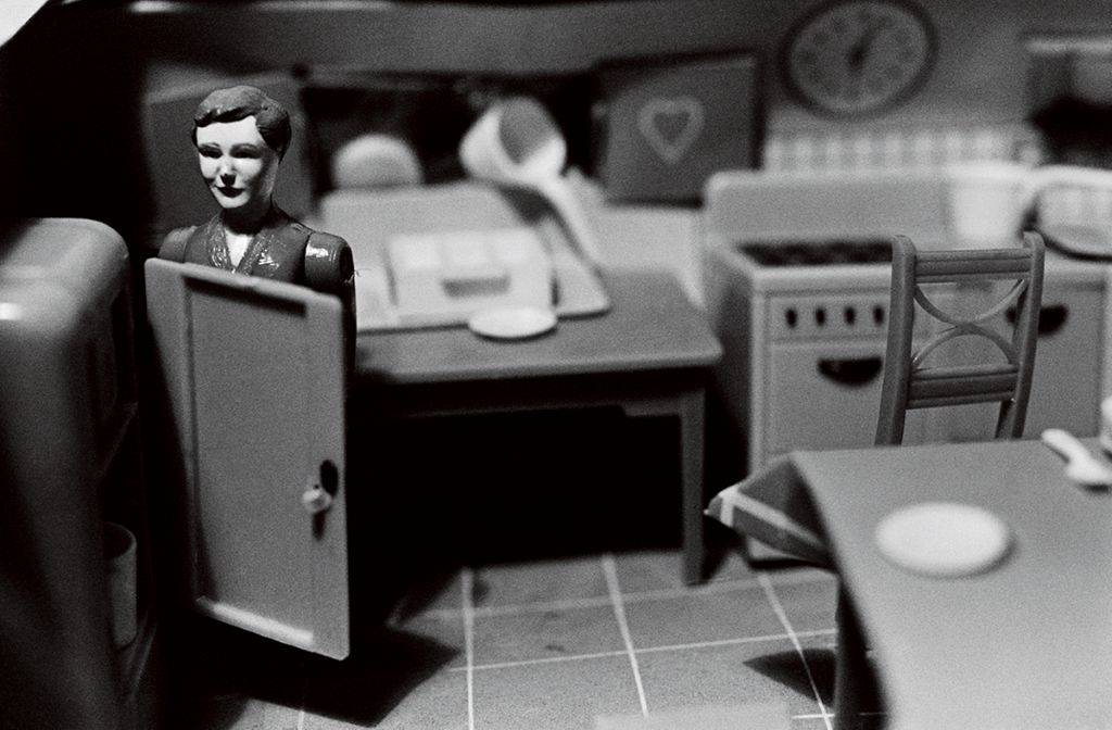 The black-and-white photograph shows a doll figure in a dollhouse kitchen. The image is out of focus, as the focus is on the doll figure standing behind the door of an open miniature refrigerator.