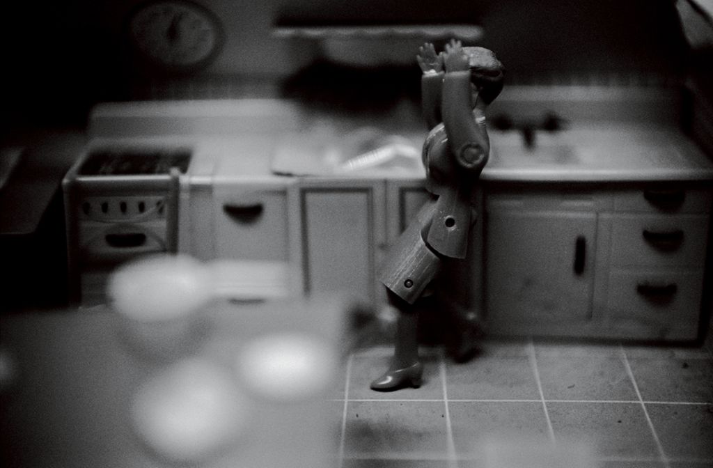 The black-and-white photograph shows a dollhouse kitchen in which a doll figure wearing a suit stands. In the foreground there is a set kitchen table, and in the background a kitchen unit. Both planes are heavily out of focus. The entire focus of the image lies in the centre, where the doll figure—its articulated limbs suggesting movement—appears to be in the process of falling.