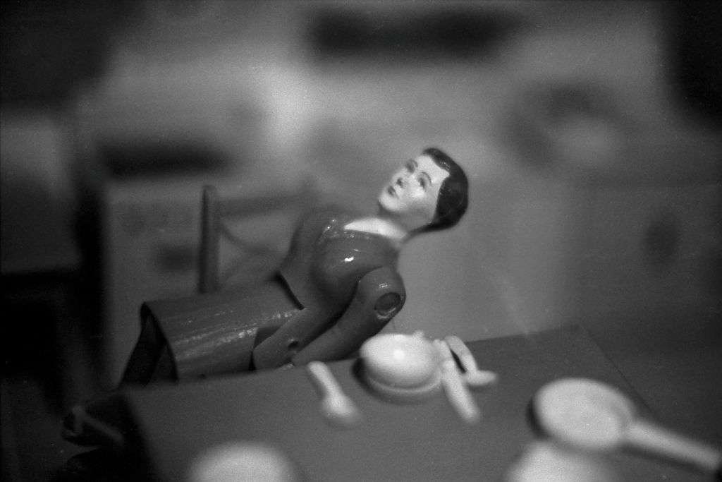 The black-and-white photograph shows a dollhouse kitchen in which a doll figure wearing a suit sits on a chair. In the foreground there is a set kitchen table, and in the background a kitchen unit. Both foreground and background planes are heavily blurred, as the focus is exclusively on the doll figure. She sits at an angle on the chair and appears to be on the verge of falling.