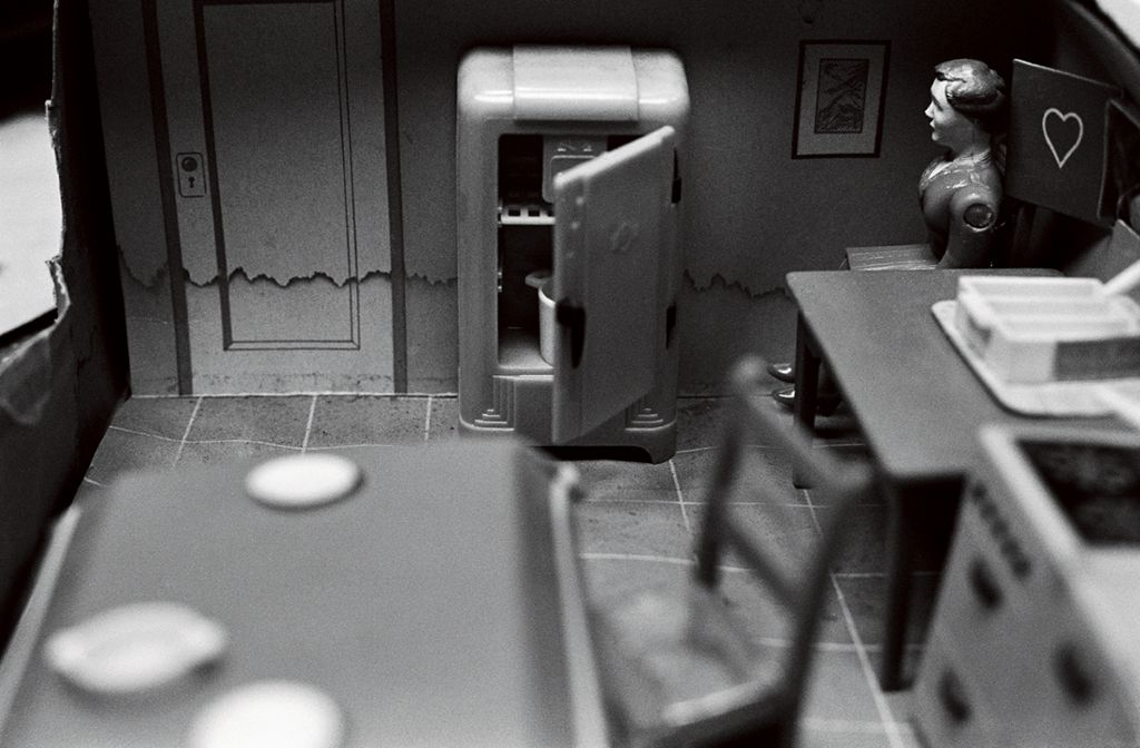 The black-and-white photograph shows a miniature dollhouse kitchen in which, alongside a refrigerator, two large tables and a kitchen unit are arranged. A doll figure sits in the background plane between the refrigerator and one of the tables. The table in the foreground is heavily out of focus.