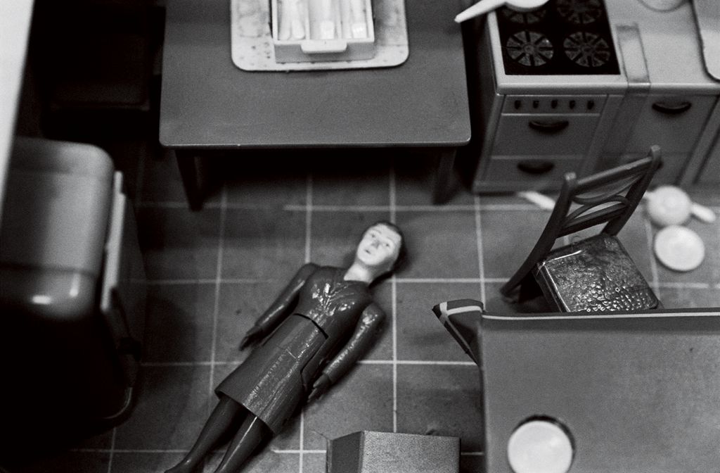 The black-and-white photograph shows a dollhouse kitchen from a bird’s-eye view. A doll figure lies on the floor between a kitchen unit and two tables. Cutlery is also scattered across the floor.