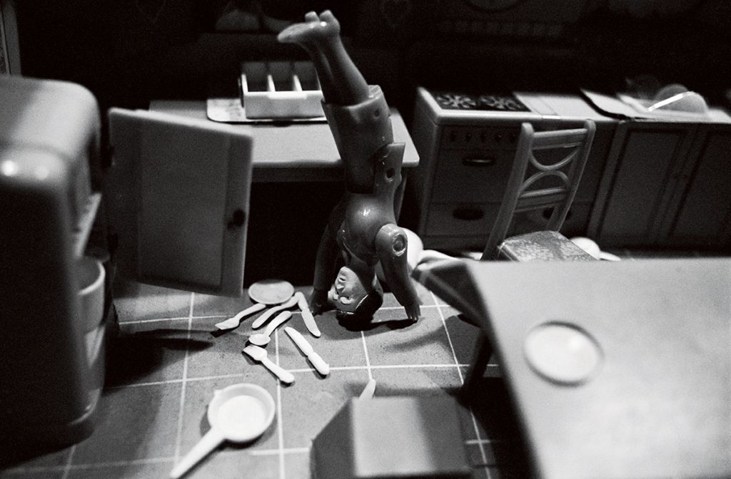 The black-and-white photograph shows a dollhouse kitchen, taken from a slightly elevated angle. The refrigerator door is open, and dishes and cutlery are scattered across the floor. Amid the mess between the kitchen unit and the dining table, a doll figure is doing a handstand.