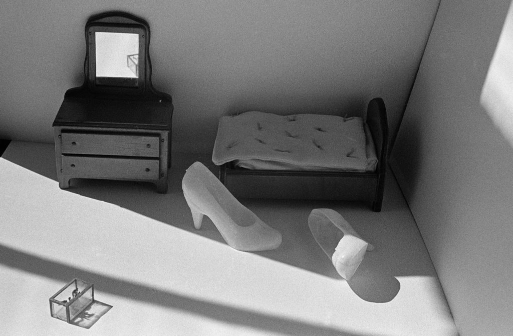 A pair of Barbie toy shoes is photographed in front of a miniature bed, next to which stands a miniature chest of drawers. At the lower left edge of the frame, a small box is placed. Soft light enters from the left side. The photograph is rendered in black and white.