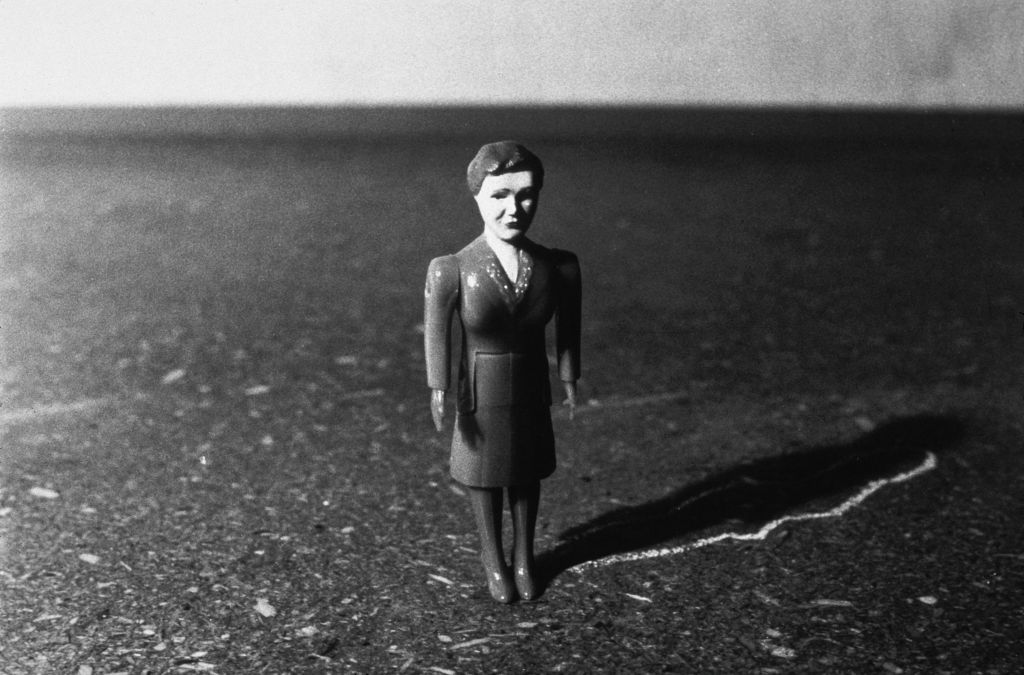 A doll figure wearing a suit stands in an unfurnished room. Light falls in from the side. The outline of the figure’s shadow has been traced on the floor with chalk.