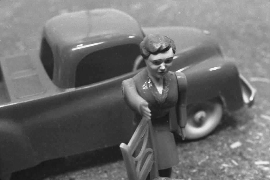 The black-and-white photograph shows a miniature doll in front of a toy car. The doll is holding a chair.