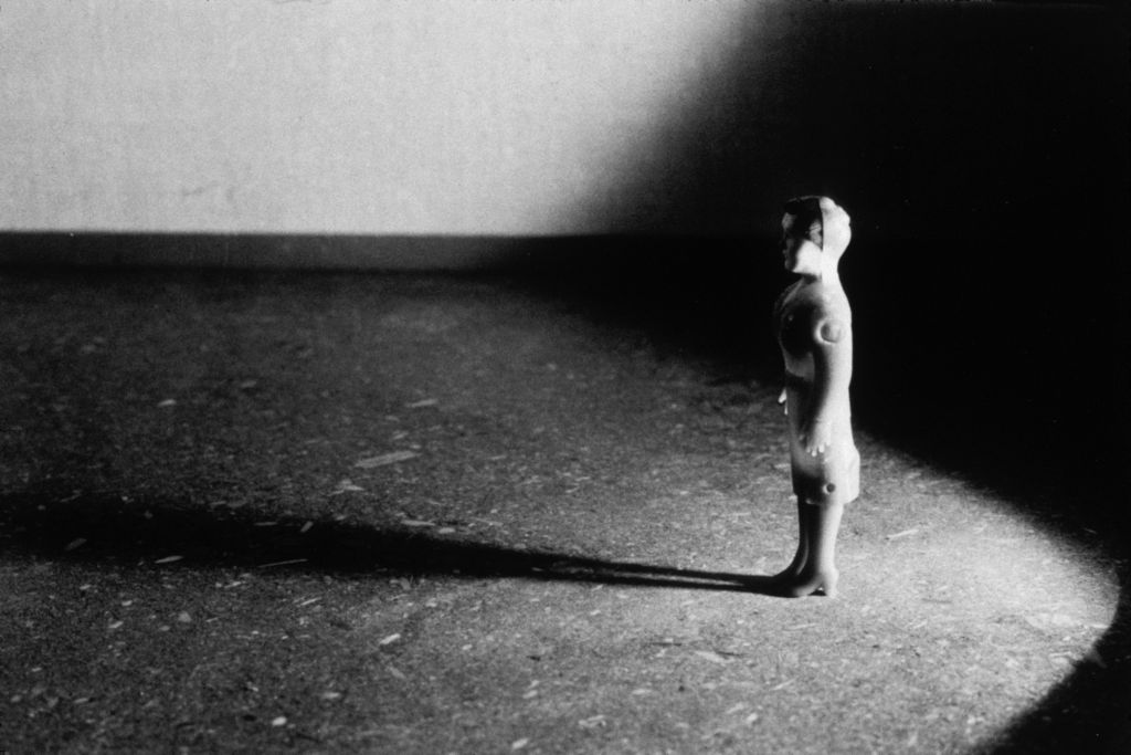 A female doll figure wearing high heels and a skirt stands in an empty room. The camera captures her in profile as she gazes straight ahead. A strong shadow dominates the space. The photograph is rendered in black and white.