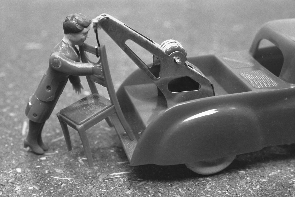 The black-and-white photograph shows a miniature doll in front of a toy car. A crane is attached to the toy car. The doll appears to be struggling to attach a chair to the crane.