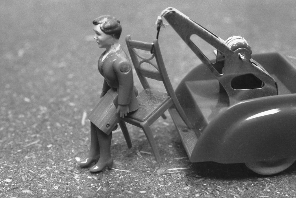 The black-and-white photograph shows a toy doll lying in front of a toy car. The toy car has a crane mounted on its rear platform. A chair is attached to the crane. It appears as though the doll lying on the ground has fallen from the toy chair.