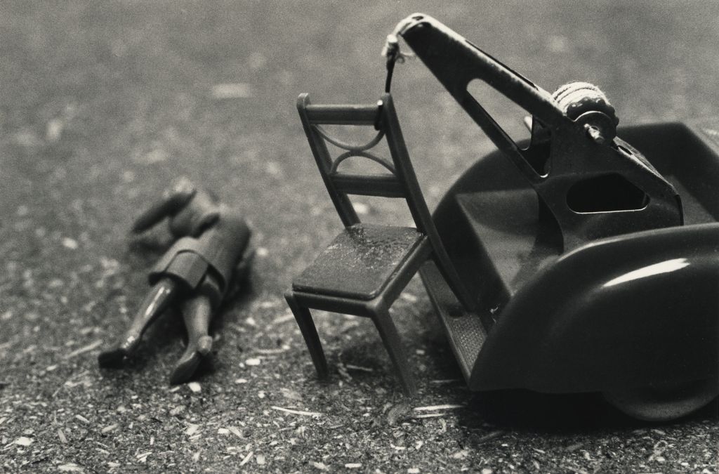 The black-and-white photograph shows a toy doll lying in front of a toy car. The toy car has a crane mounted on its rear platform. A chair is attached to the crane. It appears as though the doll lying on the ground has fallen from the toy chair.