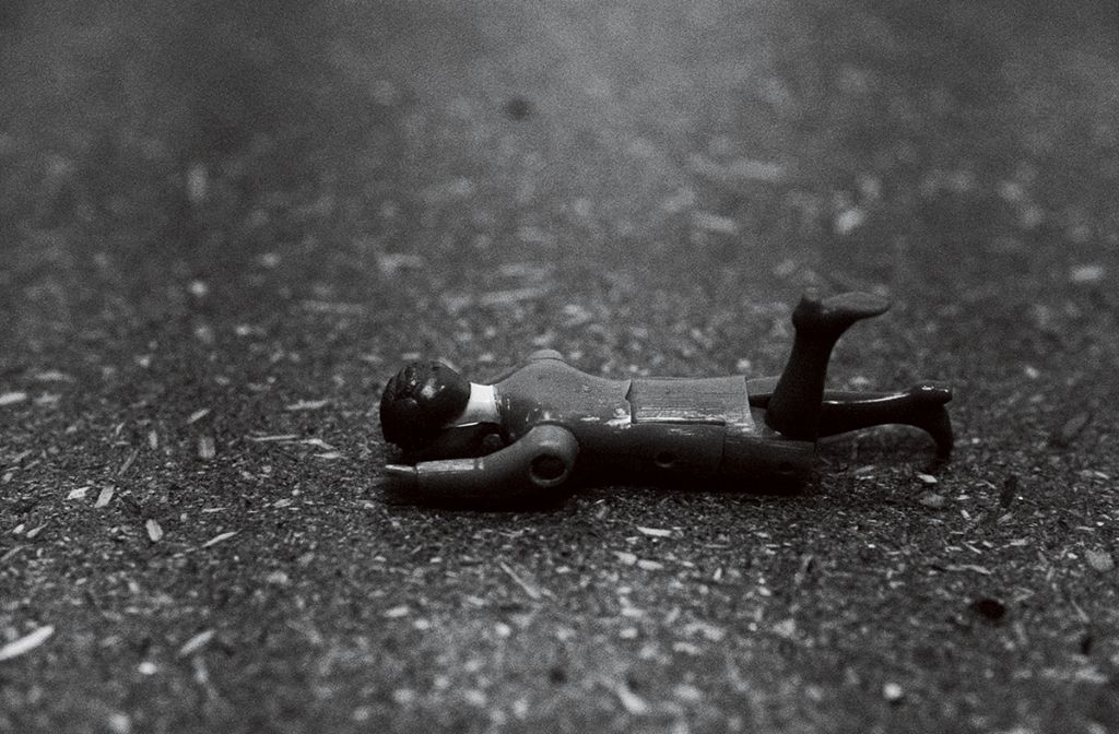 The black-and-white photograph shows a fallen toy doll. It lies in the centre of the frame with its head resting on the ground. Its left leg is slightly bent.