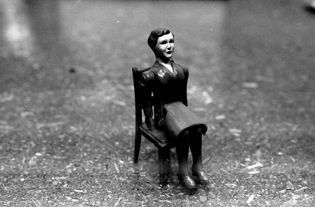 The black-and-white photograph shows a female toy figure seated on a doll’s chair in close view. The background is slightly out of focus, as the focus is exclusively on the doll.