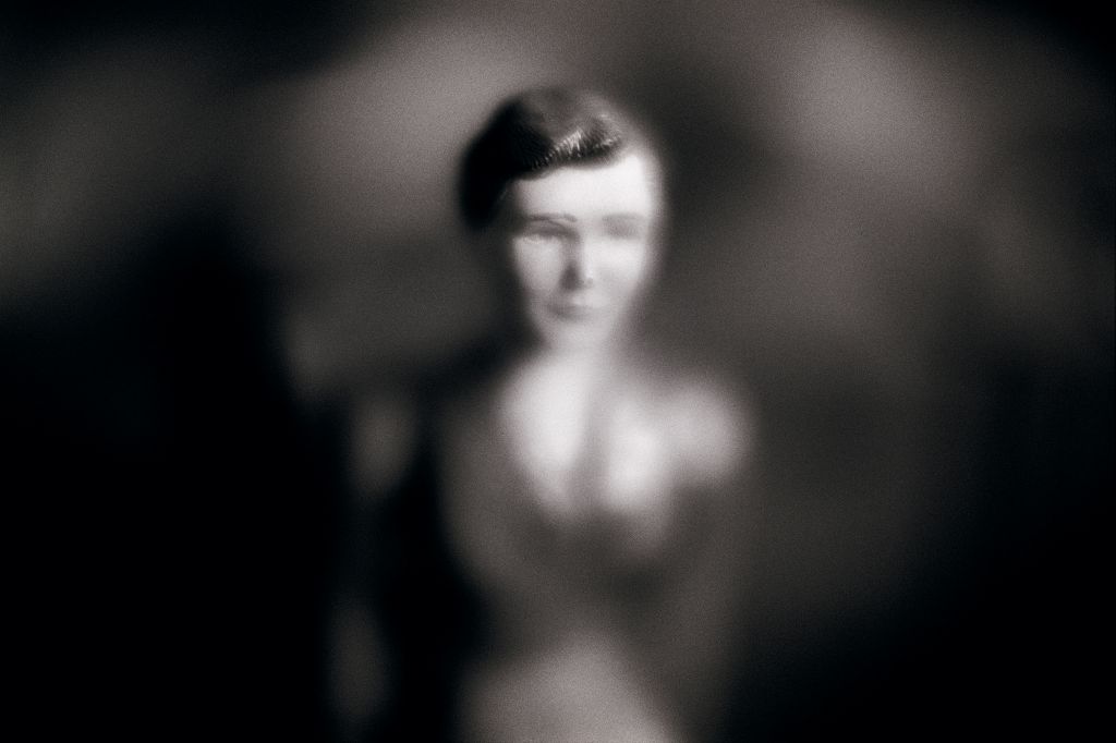 The head of a doll appears through a blurred vignette. The photograph is in black and white.