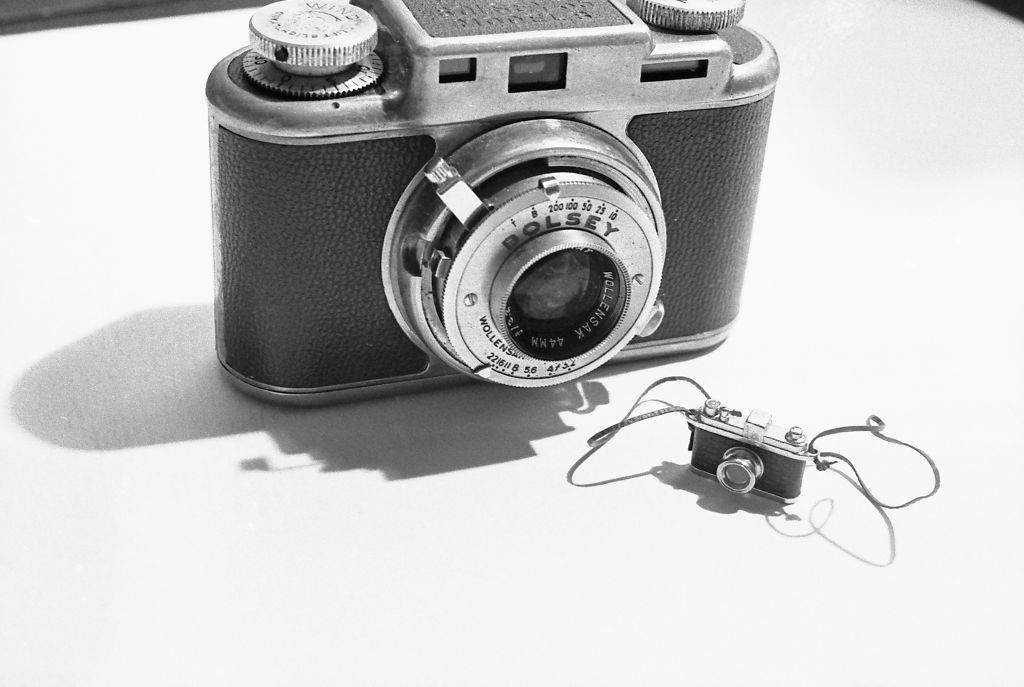 The photograph shows a black-and-white image of a miniature camera, behind which stands a full-sized camera. The photograph is rendered in black and white.