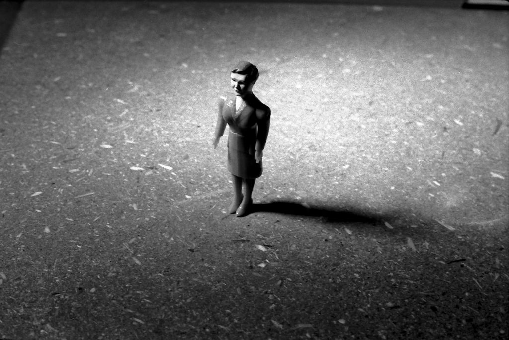 A female doll figure wearing high heels and a suit stands in an empty room. The camera captures her from an elevated, slightly left-hand angle while she gazes straight ahead. A strong shadow defines the space. The photograph is rendered in black and white.