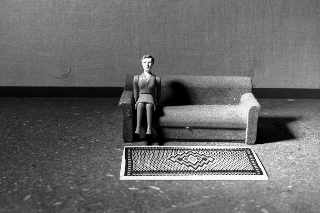 A female doll figure wearing a suit sits on a miniature sofa. She is positioned right at the outer edge of the seat. In front of her lies a patterned woven rug. The photograph is rendered in black and white.