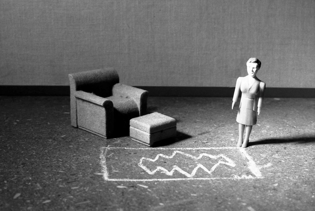 A female doll figure stands in a simple room. She stands in front of a rug drawn in white chalk. Next to the rug, an armchair with an ottoman is positioned at an angle. The photograph is rendered in black and white.