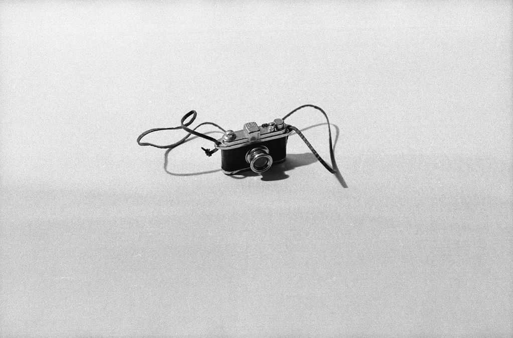 A miniature camera stands against a white background. The light falls from behind the object, casting a small shadow forward. The photograph is rendered in black and white