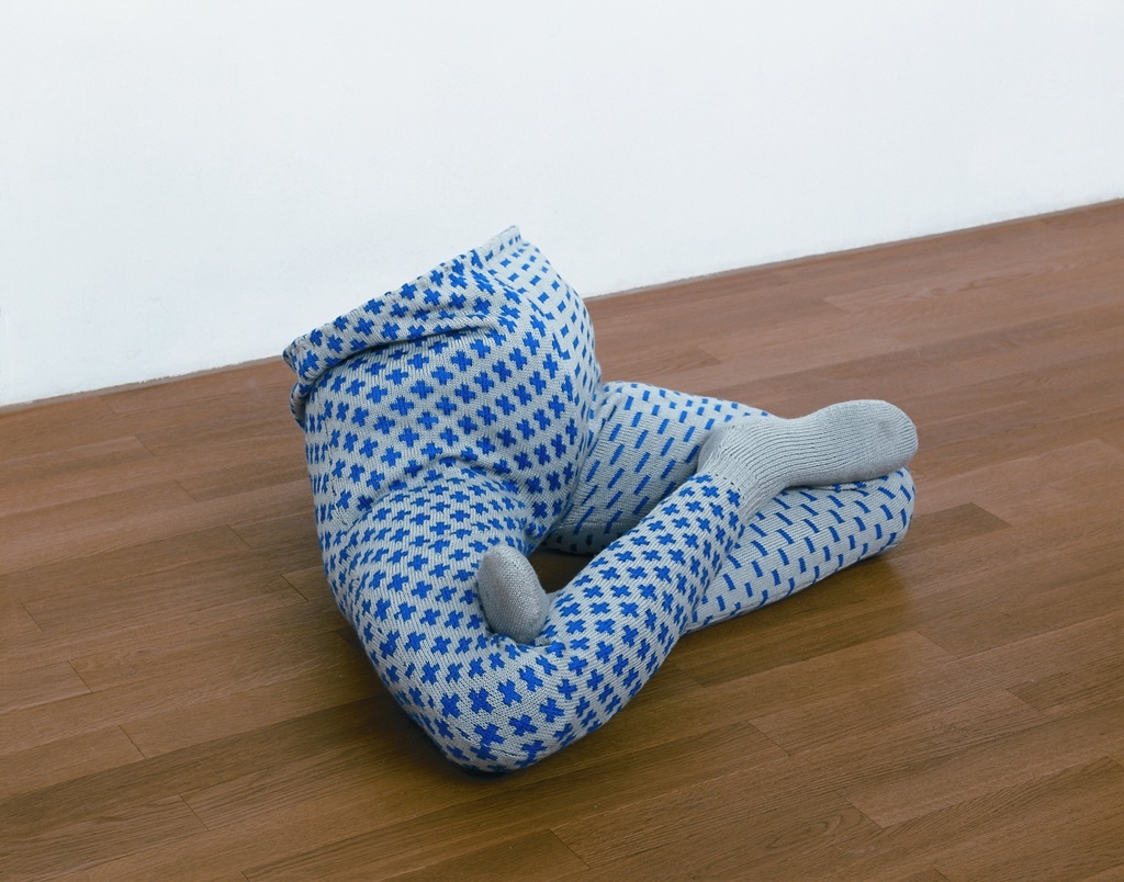 Skulptur eines Körperunterteils ohne Oberteil, im Schneidersitz auf dem Boden sitzend; die Beine tragen blau-grau gemusterte Strumpfhosen, das eine Bein trägt ein Plus-Muster, das andere ein Minus-Muster.