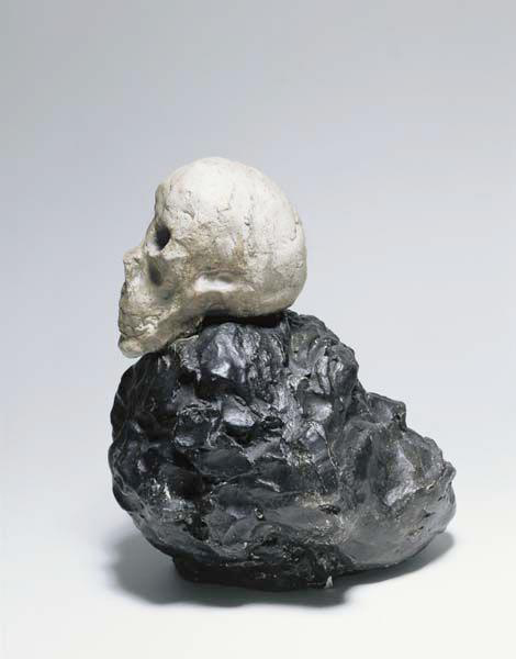 Knetförmig geformte Plastik zweier Köpfe. Der untere Kopf hat eine schwarze Farbe und ähnelt einem Affenschädel, darauf angebracht ist ein weißer Kopf in Form ähnlich eines Totenschädels.