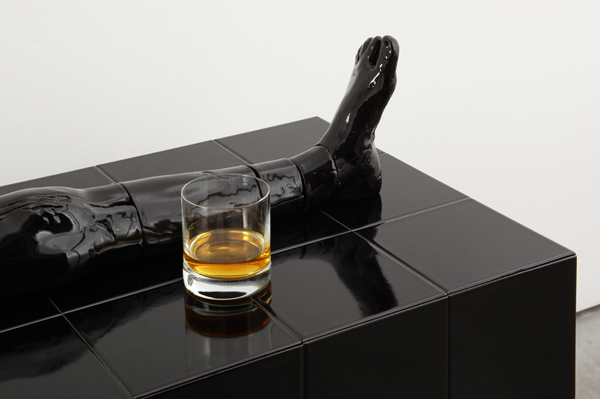  Detailaufnahme: Auf einem schwarzen hochglänzenden Tisch liegt eine schwarze Keramikskulptur, die ein Bein darstellt. Daneben steht ein Glas mit einer Spirituose.