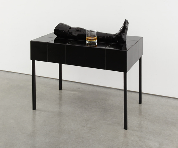 Auf einem schwarzen hochglänzenden Tisch liegt eine schwarze Keramikskulptur, die ein Bein darstellt. Daneben steht ein Glas mit einer Spirituose.