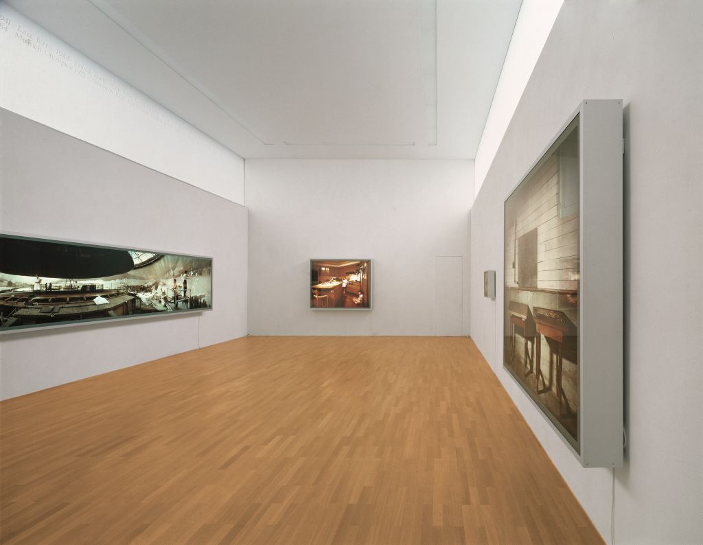 In a museum gallery, photographs by Jeff Wall are displayed on three walls. "Restoration", the work discussed here, is located on the wall to the left from the viewer's perspective.