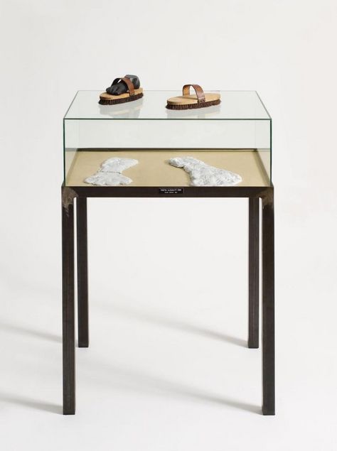 In einem Glasschaukasten mit hohen Tischbeinen liegen zwei gipsartige weiße Fußabdrücke. Auf der Glasabdeckung liegen zwei Handbürsten, in der linken steckt eine schwarze Gipsplastik eines Fußes.