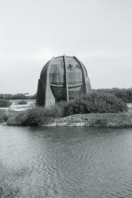 Black and white photograph of a large round concrete structure in the middle of a river landscape.