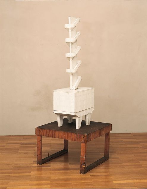 Auf einem Podest steht eine weiße Skulptur, die an ein architektonisches Modell erinnert. Ein Gebäudekorpus ist auf hohen Stützpfeilern aufgebockt, die an die Topologie von Le Corbusiers Cité Radieuse erinnern. Aus dem Baukörper ragt ein vier Geschosse hohes Treppenhaus.