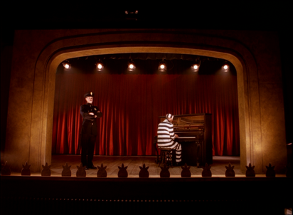 On a theater stage, a man in a black and white striped suit and cap sits at a piano with his back turned to the viewer. To the left of the piano, a man in a black uniform stands with his arms crossed, looking over the auditorium. 