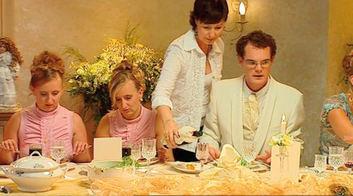 Two women with the same hairstyle and dress are sitting at a festively decorated table. Next to them sits a man in a light-colored suit, reminiscent of a groom. A woman stands behind him and pours him wine.