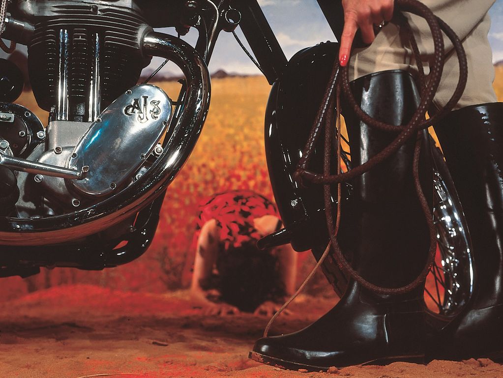 A person with high leather boots stands in front of a motorcycle, holding a long whip rolled up. You can't see the head, but judging by the hand with long fingernails and nail polish, it could be a woman. Another woman can be seen between the wheels of the motorcycle. She is kneeling on the ground, her head hanging forward, wearing a red dress with black roses printed on it. 