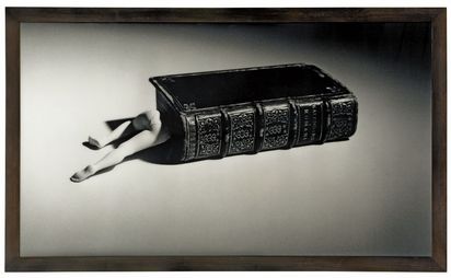 Zwei Puppenbeine ragen aus einem altmodisch in Leder gebundenen Buch hervor, das auf dem Boden liegt. Das Foto ist in Schwarz-Weiß gehalten, und das Buch wird von einem sanften Lichtkegel beleuchtet.
