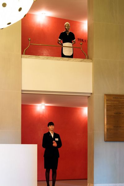 Auf einem Balkon in dem Foyer eines Opernhauses steht eine goldene Figur, die Hände hinter dem Rücken verschränkt. Die schwarz-weiße Uniform lässt vermuten, dass das Modell ein Hausmädchen darstellen soll. Auf der unteren Etage, unter dem Balkon steht eine ebenfalls schwarz weiß gekleidete Person, die zum Personal der Oper gehört.