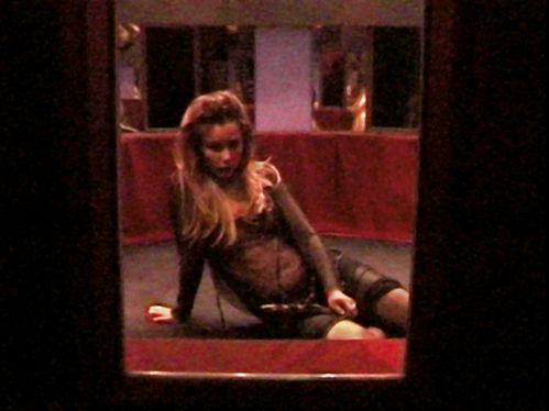 A blonde woman in black see-through clothes sits in a room furnished with red fabrics