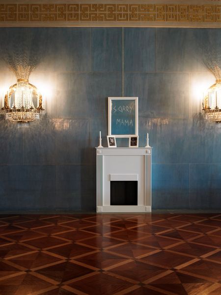 Between two candelabras stands an entirely white fireplace, set into a blue-painted wall. Above the fireplace hangs a mirror inscribed with “Sorry Mama” in angular lettering. On the mantelpiece are two black-and-white childhood photographs of Michael Elmgreen and Ingar Dragset, as well as two white candlesticks, one of which has fallen over.