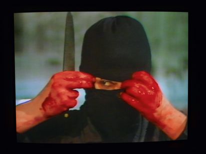 Eine Person mit schwarzer Sturmhaube und rot verschmierten Händen hält ein Messer in der Hand während sie mit einem Auge durch ein Loch in der Sturmhaube schaut.