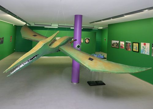 Eine Skulptur aus Pappe, Holz, Papier und Folie mit Farbe und Nägeln. Ein riesiger hellgrüner Flugsaurier hängt an durchsichtigen Schnüren von der Decke. Er hat seine langen Spitzen Flügel ausgebreitet und seine kleinen Füße, die wie Hühnerfüße aussehen, zeigen nach hinten. An seinem Hinterkopf hat er ein langes spitzes Horn, das leicht nach oben gebogen ist. Sein Kopf ist auch schmal und spitz, er hat sein Maul leicht geöffnet und darin sind viel kleine spitze weiße Zähne zu sehen. Er hat keine Augen.