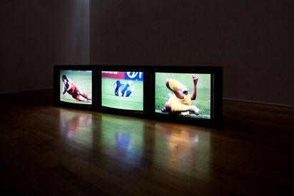 Drei Hantarex-Fernseher sind zu sehen. Auf jedem Bildschirm fällt ein Fußballspieler zu Boden. Die Spieler tragen Trikots in Rot, Gelb und Blau.