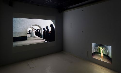 Two video projections of different sizes on one wall. The larger one shows a row of people with black hooded capes past which a woman in a wedding dress walks. The smaller one shows a naked man under a barrel vault.