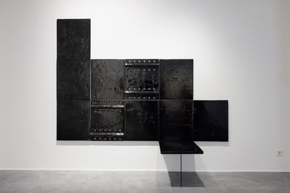 Schwarze großflächige Keramikfliesen mit einer glänzenden Lasur sind an einer Wand angebracht. Die Oberfläche ist leicht zerklüftet und aus zwei Platten sind Löcher ausgestanzt. Eine der Platten springt aus dem schwarzen flächigem Keramikobjekt hervor. Die hervorstehende Platte erweckt den Eindruck einer Sitzfläche, und wird von einem filigranem Stab gestützt. 