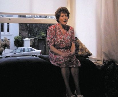 Ältere Frau in einem floralen rosa Kleid sitzt auf der Rückenlehne eines dunklen Sofas vor einem großen Fenster, Hände im Schoß, Mund geöffnet, im Hintergrund Straße mit geparktem Auto und Hausfassade.
