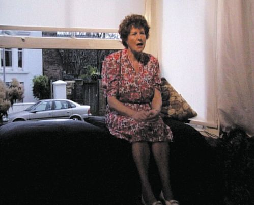 An older woman in a floral pink dress sitting on a dark sofa by a large window, hands clasped on her lap and mouth open as if speaking; a patterned cushion beside her and a silver car and residential street visible outside the window.
