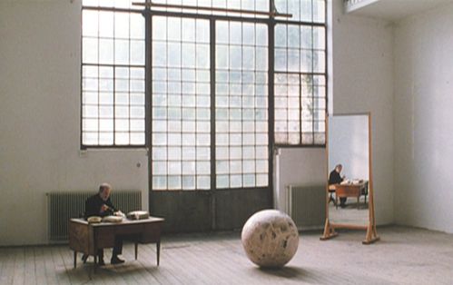 A man dressed in black is sitting at a desk in a large room with a high window wall. To his right is a large sphere on the floor, next to it a mirror that reflects the man at the desk. 