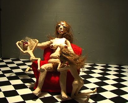 In einem Raum mit schwarz-weiß kariertem Boden sitzt die Knetfigur einer nackten weißen Frau auf einem roten Sessel. Links und und rechts von ihr sitzen zwei langhaarige, nackte Knetfigur-Mädchen. Das linke Kind sitzt auf der Armlehne beugt sich mit gehobenen Armen nach hinten, den Mund weit offen, als würde es schreien. Das rechte Kind kniet am Boden vor dem Sessel, den Rücken zum Betrachter, als würde es die Frau anflehen. Die Frau blickt mit runtergezogenen Mundwinkeln vor sich hin. 