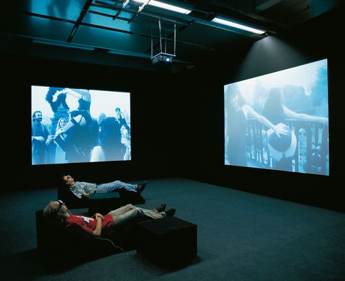 Dark projection room with two video screens with images of revealingly dressed people. Two viewers are lying on chaises longues wearing glasses