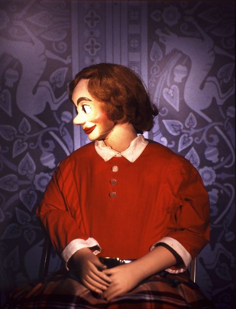 A ventriloquist dummy sits on a  chair in front of purple wallpaper with Art Nouveau ornamentation. It has particularly large eyes and a striking red mouth. Its gaze is directed slightly to the side, and it is smiling. The hair is side-parted, softly curled, and wavy. Lines around the mouth suggest that the jaw is movable. It wears an old-fashioned red shirt with a white collar and a tartan skirt.