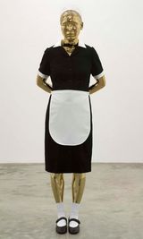 Eine goldfarbene Puppe steht vor einem neutralen Hintergrund. Sie hat die Hände hinter dem Rücken verschränkt und trägt eine Hausmädchenuniform mit einem schwarzen Kleid, einer weißen Schürze, weißen Socken und schwarzen Schuhen. Ihr Blick ist leicht nach unten gerichtet.