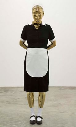 Eine goldfarbene Puppe steht vor einem neutralen Hintergrund. Sie hat die Hände hinter dem Rücken verschränkt und trägt eine Hausmädchenuniform mit einem schwarzen Kleid, einer weißen Schürze, weißen Socken und schwarzen Schuhen. Ihr Blick ist leicht nach unten gerichtet.